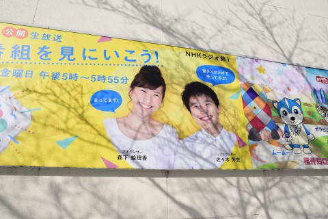 two broadcasters (a male and a female) are in the poster of NHK Fukui on the outside wall of their building 